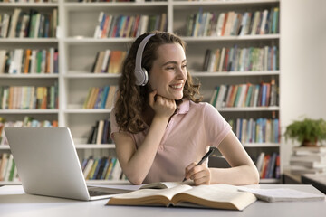 Fototapeta Happy college student girl in wireless headphones enjoying studying in library, sitting at laptop, open books, working on essay, article, writing notes, looking away, thinking, smiling, laughing obraz