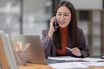 Cheerful business Asian woman freelancer wear glasses making telephone call share good news about project working in office workplace, business finance concept.