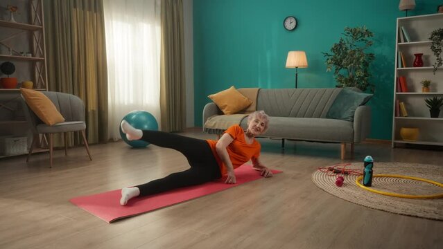 An elderly woman lies on a sports mat on the floor in the living room and does sports. The woman lies on her side, leaning on her elbow and swinging her leg. Concept of sportive lifestyle, wellness.