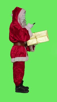 Vertical video Profile of santa claus taking picture of gift box over full body greenscreen backdrop, checking present before delivering to kids. Young man saint nick character with smartphone and