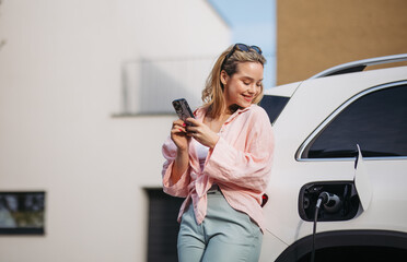 Young woman with smartphone charging her electric car in home, sustainable and economic...