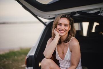 Young woman sitting in a car trunk near lake, enjoying summer time.