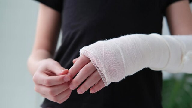 Child doing massage on fingers of broken hand wrapped in white plaster bandage.  Concept of healthcare. The girl holds a broken arm against the background of a black t-shirt