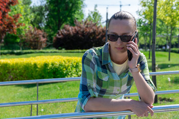 Fototapeta premium Woman stands near metal railings and talking on mobile phone. Young adult use smart phone for chatting. Female resting and conversation on cell phone. Leisure, technology and modern lifestyle concept.