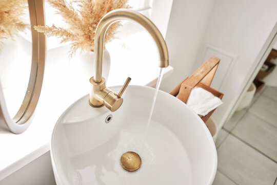 A beautiful sink with a golden faucet next to an oval mirror and a shelf with hand towels. Close-up of an elegant golden faucet in the bathroom sink next to stylish decorations.