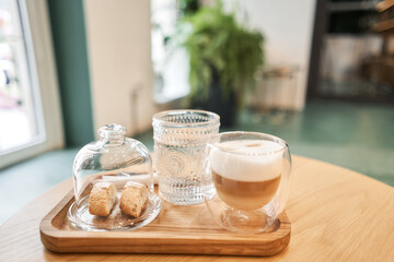 Cup of coffee cappuccino with cantuccini and a glass of water on a wooden tray. Coffee with milk, a cookie and a glass with water on a glass table.