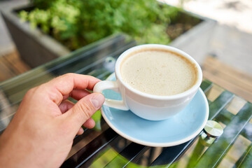 A man drinks coffee, holds a cup of hot Peanut latte in his hand. The table in the coworking space of the coffee shop. A cup of coffee on the veranda on a sunny day. Coffee break at the cafe. Close-up