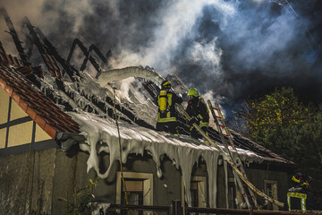 Zwei Einsatzkräfte der Feuerwehr bekämpfen einen Dachstuhlbrand mit Löschschaum. Rauch steigt...