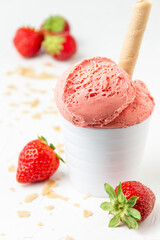 Close up of strawberry ice cream in white bowl on white table with two strawberries, wafers and crumbles, selective focus, vertical