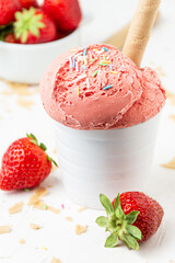 Close-up of strawberry ice cream in white bowl with wafer on white table with strawberries and crumbs, selective focus, vertical