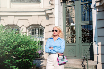 A young business lady dressed in a suit is standing in a big city. Blonde hair, blue shirt, white trousers, sunglasses.a woman stands outside the office outdoors in summer, warm weather