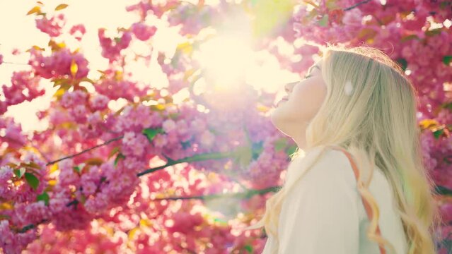 Happy blonde hair woman back rear view wind blow petals fall pink sakura flowers tree branch park summer garden sun light. girl smiling face inhales aroma flower. walk relax enjoy spring nature day 4k