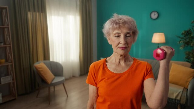 Portrait of a grayhaired elderly woman doing fitness exercises with dumbbells at home close up. A mature woman pumps up her biceps, trains her triceps, keeps her arm muscles in good shape.
