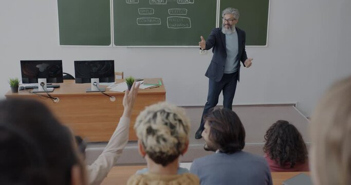 Teacher of English speaking to group of students pointing at chalk board teaching grammar rules in classroom. Language learning and education concept.