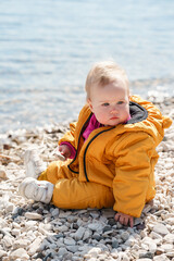 Cute baby toddler sitting on pebbles by the sea in the cold season