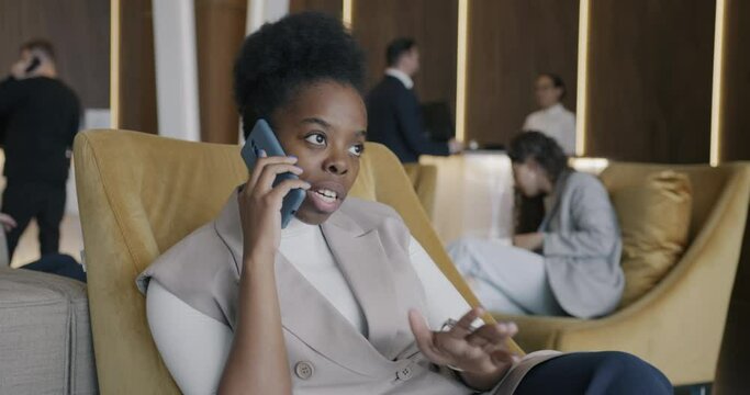 Attractive businesswoman is talking on mobile phone in hotel hall while people are busy at reception desk in background. Tourism and communication concept.