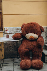 Lviv, Ukraine - May 13, 2023: big soft brown fluffy teddy bear sitting outside on chair near cafe and waiting to be served. Large teddy bear sits on chairs, beckoning visitors..