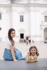 Portrait of pretty young woman with her adorable daughter sitting outdoor on stone bench