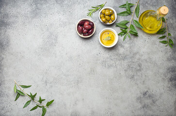 Composition with green and brown olives, extra virgin olive oil in glass bottle, olive tree branches on gray concrete stone rustic background overhead, copy space border