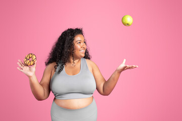 Excited african american overweight lady in sportswear tossing green apple, chooses between donut or fruit