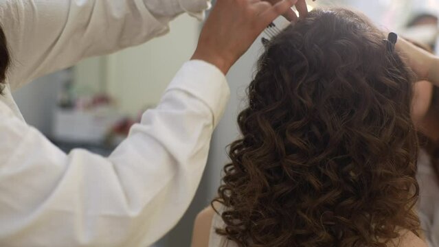 Close-up hands of unrecognizable female hairdresser styles long, curly strands of hair with comb and hands after applying of fixer in modern beauty studio. Concept of professional fashion hairstyling.