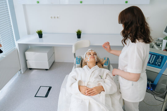 High-angle view of beautician physiotherapist consulting young woman client during facial myostimulation procedure in beauty clinic. Concept of rejuvenating skin, preventing aging.