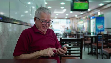 One happy elderly man using cellphone device sitting inside Brazilian cafeteria laughing at online content