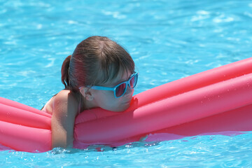 Happy child girl relaxing on inflatable air mattress in swimming pool on sunny summer day during tropical vacations. Summertime activities concept