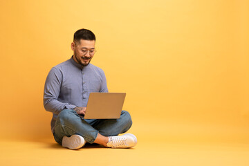 Freelance Career. Happy Asian Guy Using Laptop Computer Over Yellow Studio Background