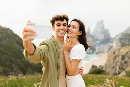 Happy european couple after engagement taking selfie on cellphone, standing on rocks cliff near ocean shore