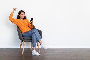 Emotional eastern woman sitting in armchair, using smartphone, copy space