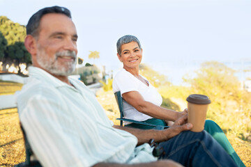 Senior Couple Sitting In Chairs Holding Coffee At Park Outdoors