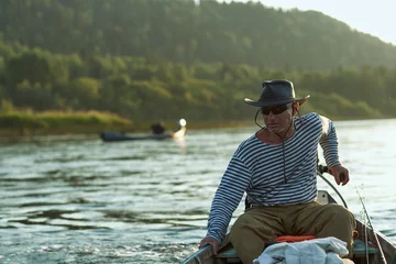 Foto op Canvas A bruA brutal man, a fisherman aged 60 plus in a vest and a black hat, drives a motor boat on the river. Sunset time on the river. The concept of tourism and recreation. High resolution photo © Marina