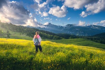 Girl on the hill with yellow flowers and green grass in beautiful alpine mountain valley at sunset in summer. Landscape with young woman in alps, rees, sky with clouds. Travel and Hiking. Slovenia - obrazy, fototapety, plakaty