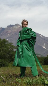 Fashionable woman waves skirts of long green cloak and covers her face with it. Stylish blonde woman posing in summer forest on background of mountain. Vertical slow motion. Part of video series
