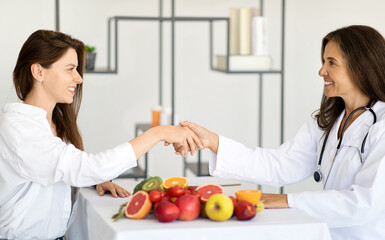 Happy adult european woman doctor nutritionist in white coat shaking hands with young lady patient
