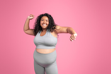 Excited african american oversize woman in sportswear dancing, celebrating success, pink background, copy space