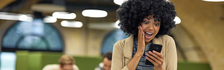 Good news. Young happy mixed race woman looking at smartphone and feeling excited while standing in coworking space