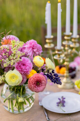 Elegant fancy wedding table decor with bright ranunculi flowers in a vase outdoors, chandelier, field or meadow. Summer, sunset, golden hour. Romantic dinner for a couple in restaurant outside