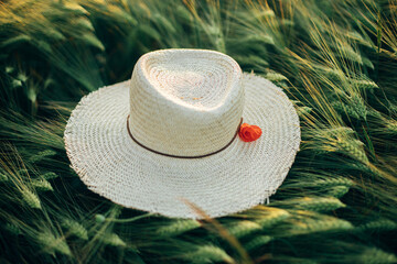 Rustic straw hat and red poppy on barley ears in evening field close up. Wildflowers and farm hat in summer countryside. Atmospheric moment in evening meadow