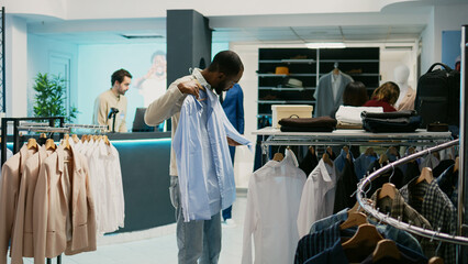 African american client examining fabric of formal shirts hanging in department store at mall. Male...