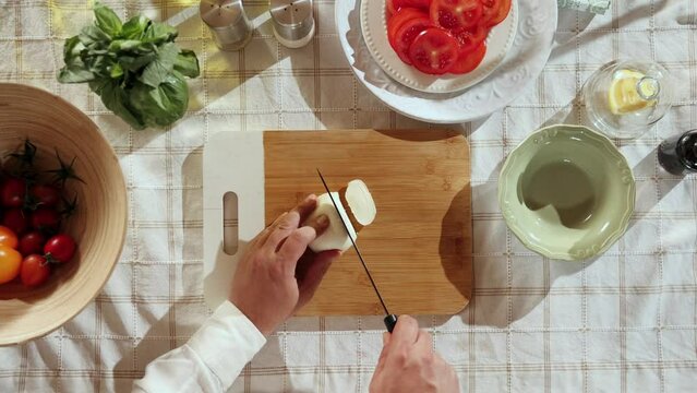Caprese salad preparation. Top view image of man's hands cutting mozzarella cheese on cutting board in the kitchen. Concept of hobby, culinary, food preparation, taste, diet, healthy dish, ad
