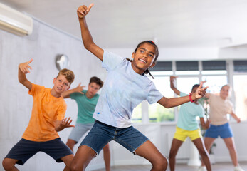 African boy performs movements during warm-up, limbering-up part of workout together with peers. Group of young girls and guys dance modern waacking in fitness club unfocused