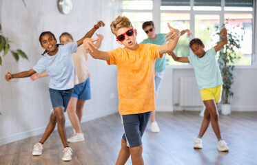Fototapeta premium Boy in sun glasses performs choreographic exercises and teaches energetic mobile social dance Jazz-modern together with friends. Multi-racial group of children performs movements in spacious studio