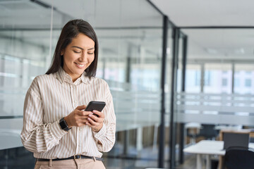 Young happy smiling professional Asian business woman manager, female worker holding cellphone using mobile phone standing in office hall working on smartphone texting message.