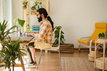 Casual adult man with prosthetic leg using laptop at desk in home office, remote work. Portrait of happy bearded guy smiling. Businessman managing internet business at home