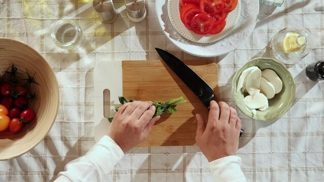 Caprese salad preparation. Top view image of man's hands cutting basil on cutting board and adding to cheese and tomatoes. Concept of hobby, culinary, food preparation, taste, diet, healthy dish, ad