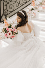 portrait of a young girl from the back, with beautiful hair and makeup in a luxurious wedding dress on the steps of a restaurant near rose flowers. Vertical photo.