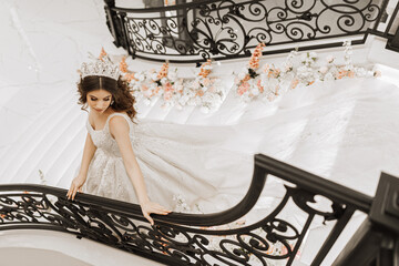 Beautiful bride in a white dress with a long train and a crown on her head in a chic interior, going down the stairs © Vasil