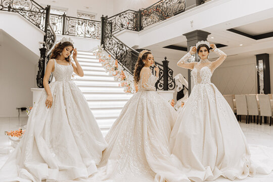 Three beautiful girls in luxurious wedding dresses with long trains pose in the hall near the steps of a restaurant decorated with flowers. Photo session for a wedding salon. Stylish fashion